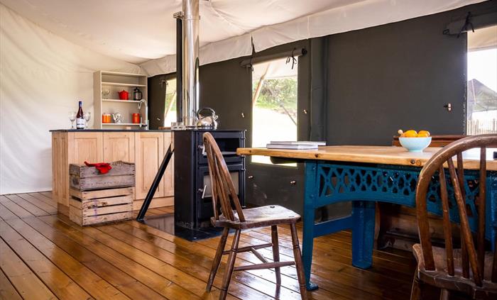 Enfys dining room, showing a big table with teal base, 2 chairs, a wood burner with oven and a slate and oak kitchen unit in the background